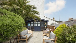 A cozy outdoor seating area with two wooden chairs and a table, a small wooden structure with a dark door, and lush greenery surrounding the space. - St Clements Cottage - Free WiFi - Outdoor kitchen and shower - Wood Burner (Mousehole)