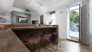 A kitchen with a wooden countertop, bar stools, a coffee machine, and a view of a garden through a glass door. - St Clements Cottage - Free WiFi - Outdoor kitchen and shower - Wood Burner (Mousehole)