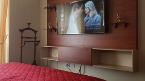 Et hotelværelse med en seng, et ophængt fjernsyn der viser et program, en træstol og en hylde.