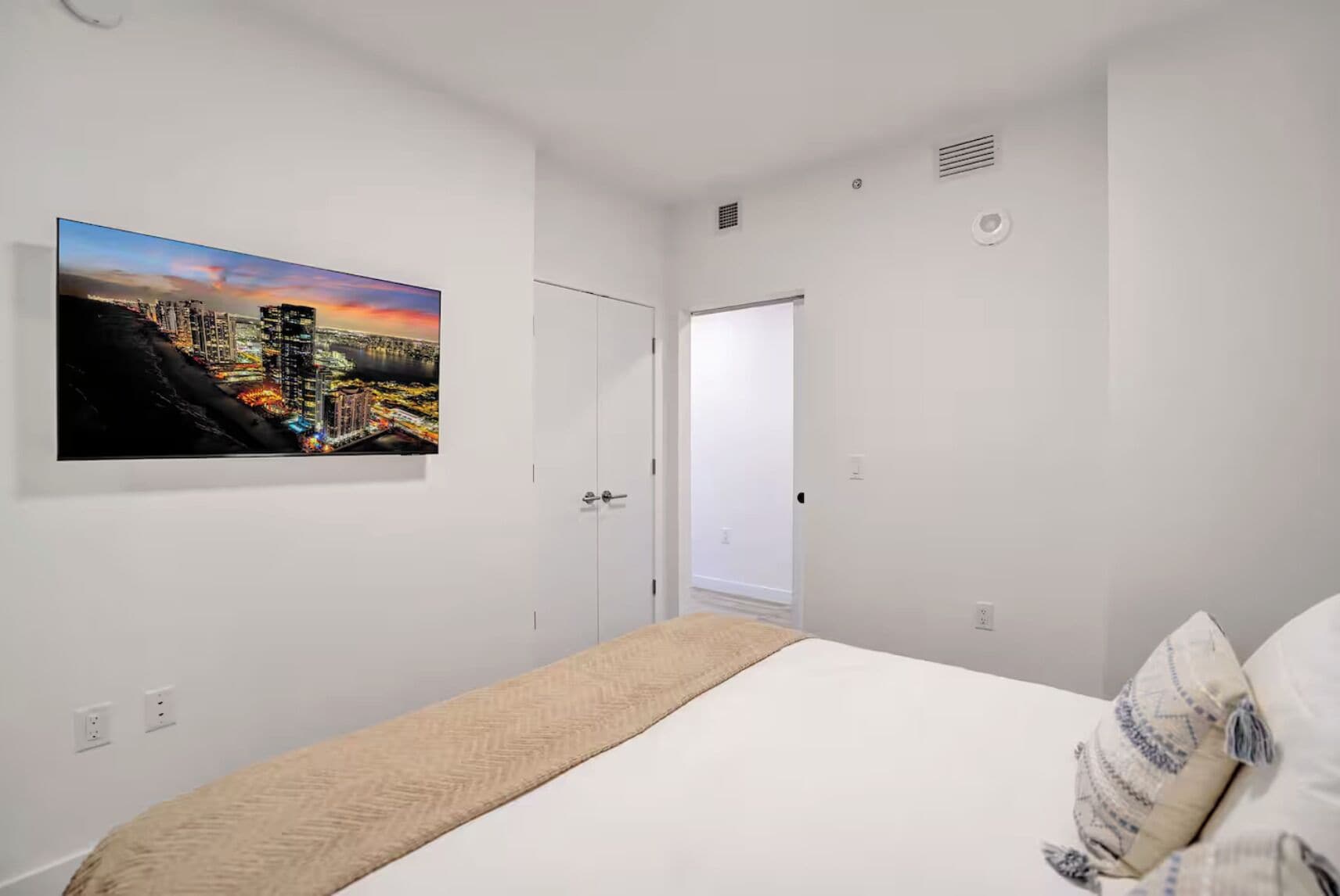 A bedroom with a bed, a door, a window, and a cityscape painting on the wall.