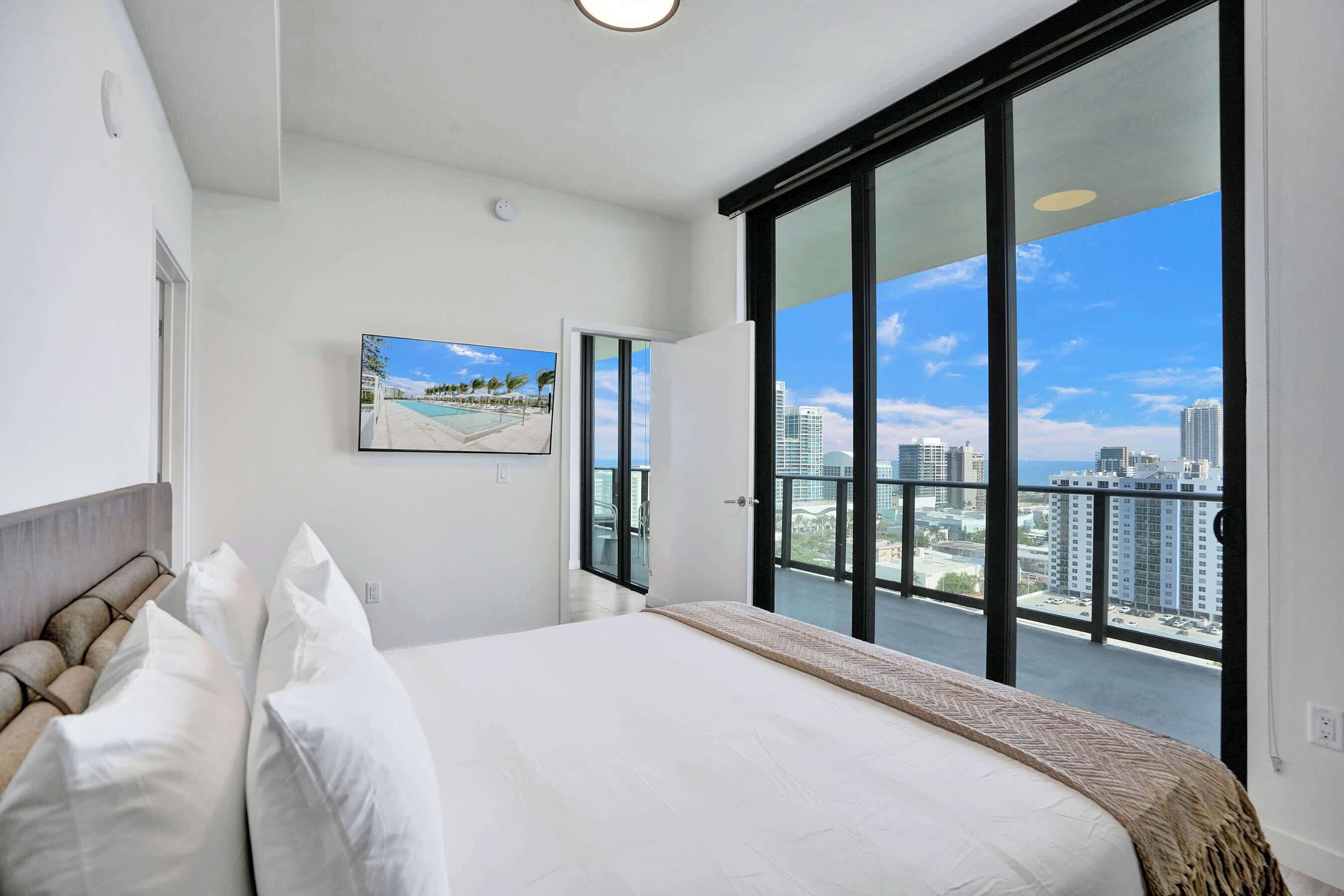 Une chambre moderne avec un grand lit, une vue sur la ville grâce à de grandes fenêtres du sol au plafond et un tableau accroché au mur.