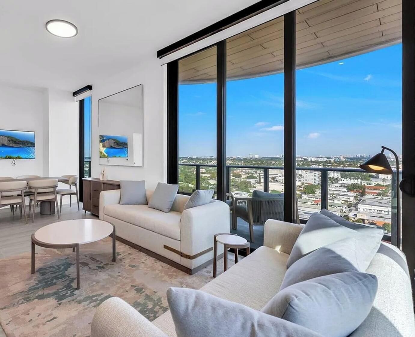 A modern living room with a sofa, coffee table, and TV, large windows with a city view, and a balcony with a lamp.