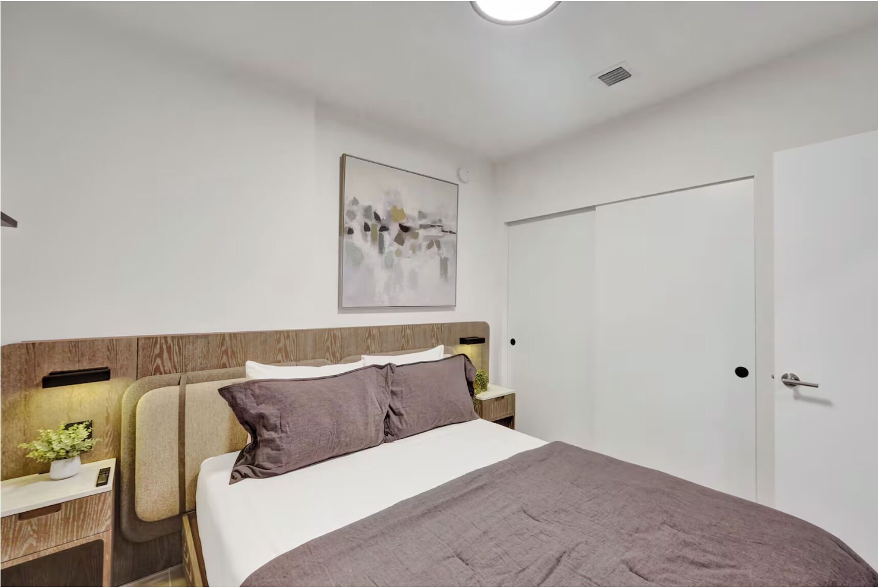 A bedroom with a bed, a nightstand, a wall-mounted light, a door, and a painting.