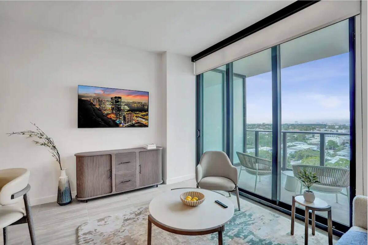 Una sala de estar moderna con un ventanal grande, un televisor de pantalla plana, un mueble gris y una mesa de centro redonda.