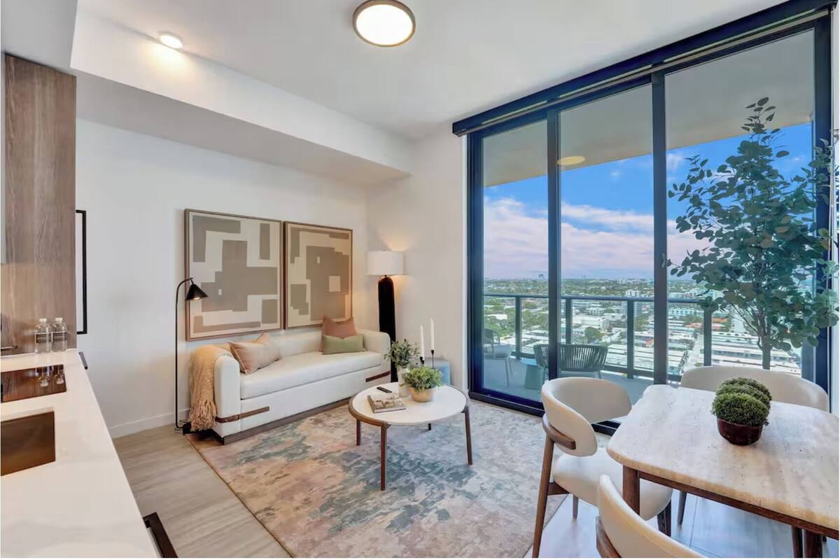 Una sala de estar moderna con sofá, mesa de centro y sillones, un comedor con mesa, y un amplio ventanal con vistas a la ciudad.