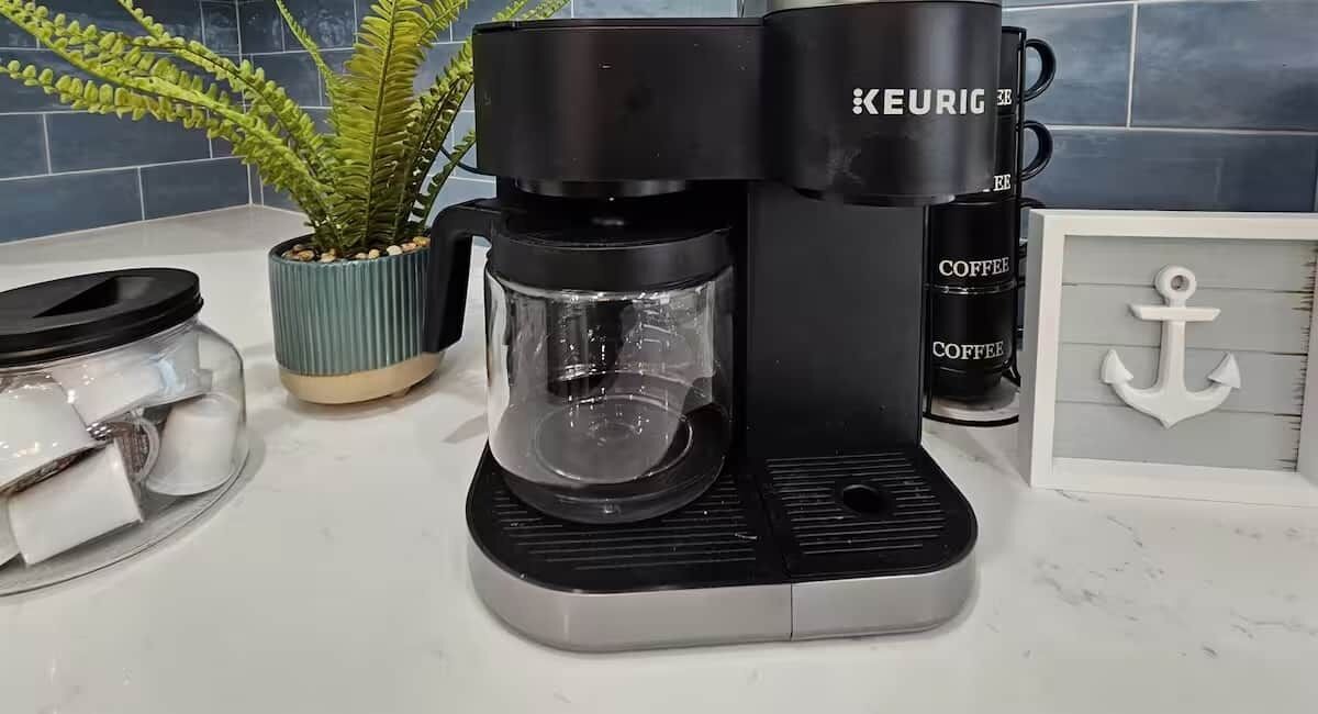 A Keurig coffee maker with a glass carafe, a potted plant, a jar of sugar, and a framed anchor decoration on a kitchen counter.