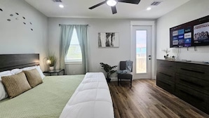 Una habitación de hotel con cama, ventilador de techo, televisor, planta y ventana con cortinas.