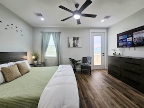 A hotel room with a bed, a ceiling fan, a TV, a plant, and a window with curtains. - Blue Turtle Inn - Sleeps 16, EV Charger, near the beach! (Bolivar Peninsula)