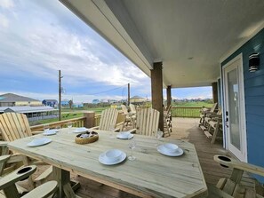 A covered outdoor deck with a dining table set for four, surrounded by wooden chairs, overlooking a grassy area. - Blue Turtle Inn - Sleeps 16, EV Charger, near the beach! (Bolivar Peninsula)