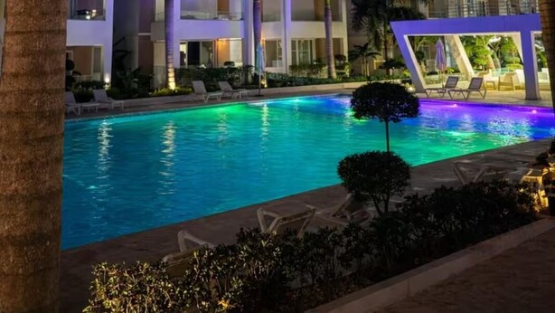 Una piscina con un elemento acuático geométrico, rodeada de palmeras y edificios modernos de noche.