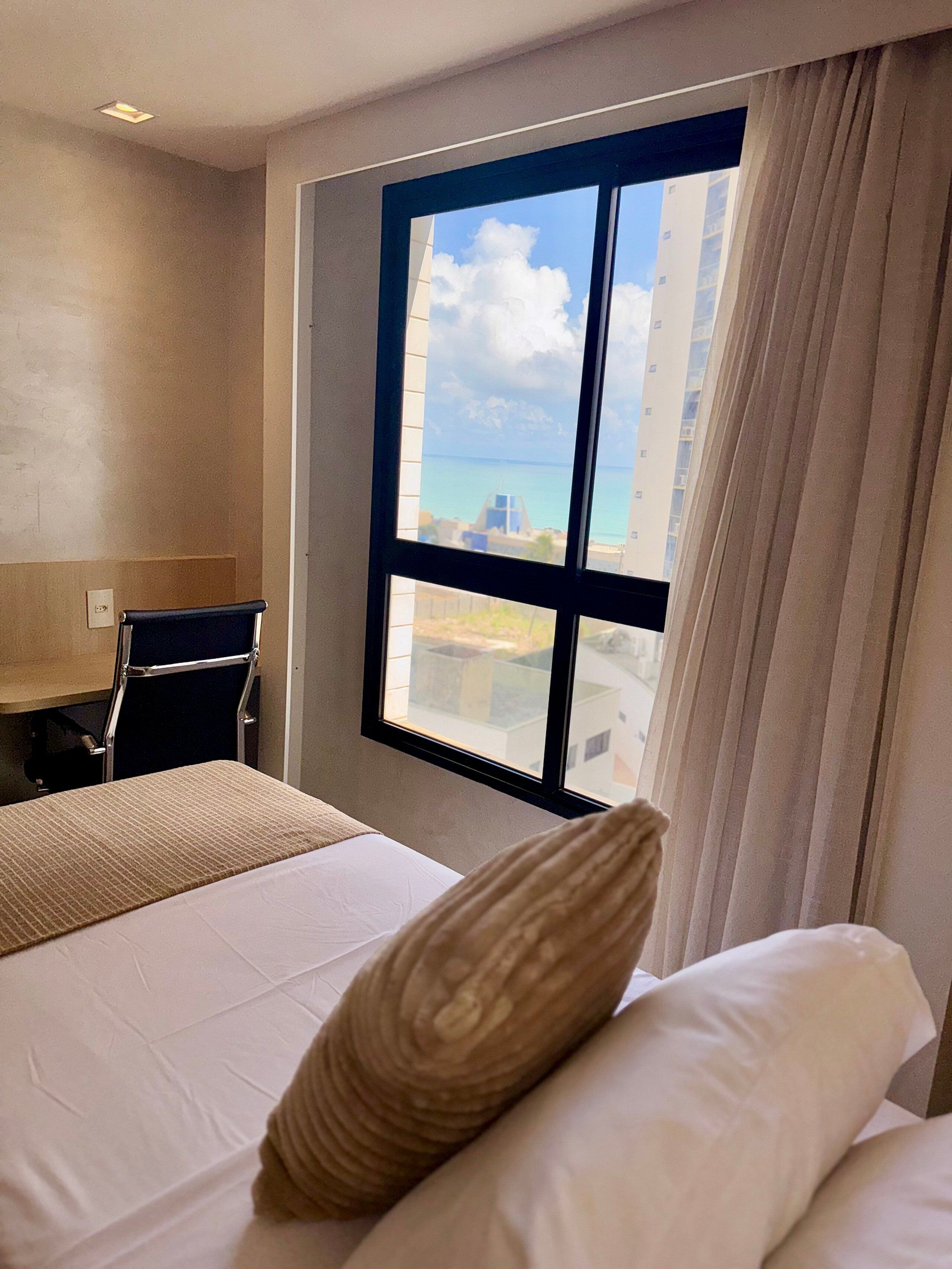 Habitación de hotel con cama, escritorio y ventana con vistas al mar y edificios.