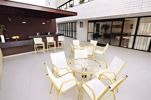 Zona salotto esterna moderna con tavoli rotondi e sedie bianche, un bancone bar e un'ampia finestra che offre vista sull'interno.