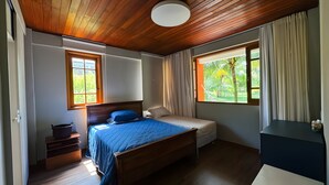 A bedroom with a wooden ceiling, a bed with a blue bedspread, a bedside table, a chair, and a window with curtains. - Village with 2 Suites Facing Timeantube Lagoon - Timeantube Reserve, Praia do Forte (Açu da Torre)
