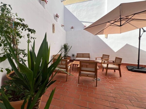 An outdoor patio with terracotta tiles, wooden furniture, potted plants, and a large umbrella.
