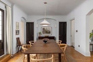 A dining room with a long wooden table, wicker chairs, a hanging light fixture, and framed artwork on the walls.