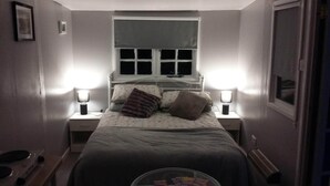 Ein schmales Zimmer mit einem Bett, zwei Nachttischlampen, einem Fenster und einer Heizung.