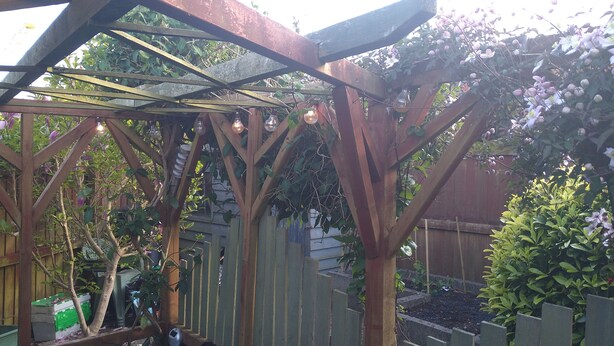 Eine Pergola aus Holz mit Dach, umgeben von Grünpflanzen und einem Holzzaun.