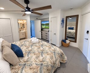 Une chambre avec un grand lit, un ventilateur de plafond, une télévision, une salle de bain avec un miroir et une porte donnant sur une autre pièce.