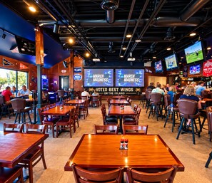 Un restaurant doté de nombreuses tables et chaises, un bar avec des téléviseurs et un grand écran diffusant des images de « MARCH MADNESS » et affichant le message « LES MEILLEURES AILLES DE POULET DE L’ÎLE, SELON UN VOTE ».