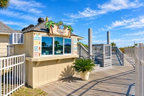 Un stand de vente de glaces situé en bord de mer, doté d’une terrasse en bois et d’un panneau publicitaire présentant les différentes saveurs de glaces proposées.