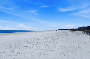 En strand med klart skys og sandet kystlinje, samt bygninger i det fjerne.