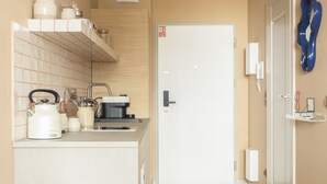 A kitchen with a white countertop, a coffee maker, and a kettle. There is a white door with a fire exit sign, a blue abstract art piece, and a small shelf with a phone and a container. - LILY APART Żurawie Premium stay (Gdańsk)