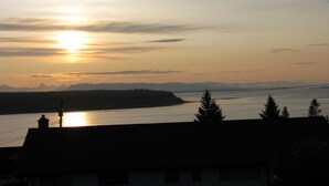 Un coucher de soleil sur une rivière, avec des montagnes à l'horizon et la silhouette d'une maison au premier plan.