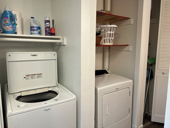 Un cuarto de lavandería con lavadora y secadora de carga frontal, repisas con útiles de limpieza y un cesto.