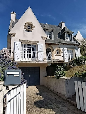 Ein großes, zweistöckiges Haus mit einem auffälligen Rundbogenfenster aus Stein, einem Balkon mit weißen Geländern und einer Garagentür.