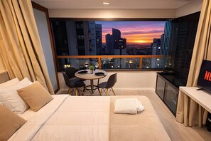 Habitación de hotel con cama, una pequeña mesa con sillas, televisor y un ventanal con vistas a la ciudad.