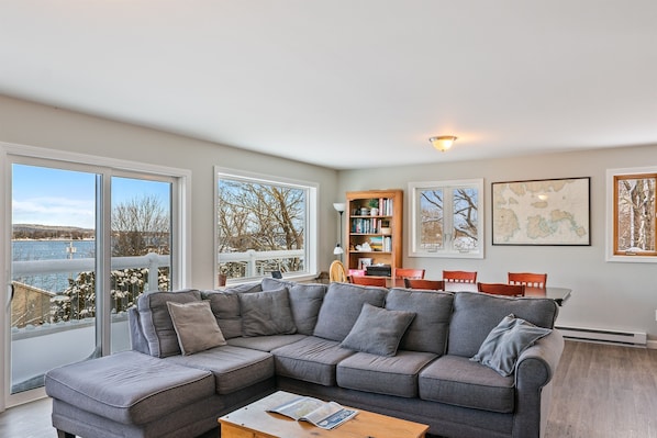 A modern living room with a sectional sofa, a dining table, and chairs. There are large windows with a view of a body of water and trees.
