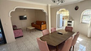 Een eetgedeelte met een houten tafel en roze stoelen, een bank en een televisie in een woonkamer.