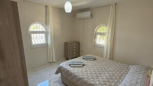 Een slaapkamer met een bed, twee ramen, een airconditioning, een houten garderobe en een nachtkastje met een lamp.