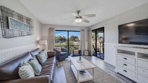 A modern living room with a sofa, armchair, coffee table, TV, and a balcony with a view. - Fairway to Shore - your peaceful, fun and cozy beach villa! (Panama City Beach)