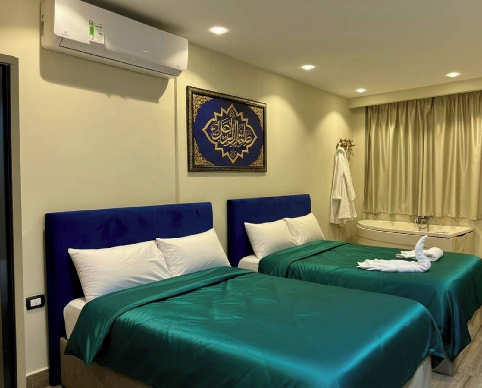 A hotel room with two beds, a wall-mounted air conditioner, a decorative wall piece, a window with curtains, and a bathroom with a towel rack.