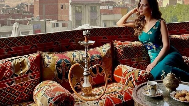 Una mujer sentada en un sofá estampado, con una pipa de agua y una tetera sobre una mesa, mirando un paisaje urbano con pirámides al fondo.