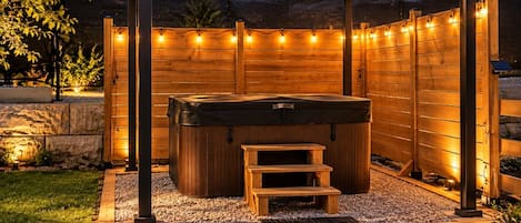 A hot tub under a wooden pergola with string lights, surrounded by a wooden fence and gravel.