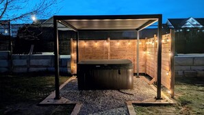 Puinen pergola, jonka alla on poreallas, ja pergolaa koristavat valonauhat. Porealtaan ympärillä on soraa ja ruohoa.