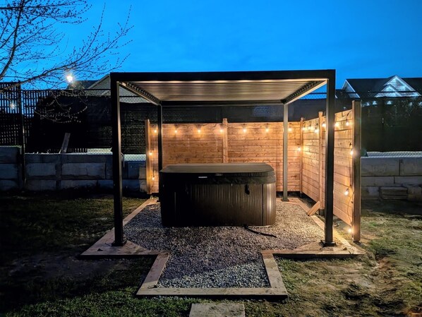 Un bain à remous sous une pergola en bois, illuminé de guirlandes lumineuses, entouré de gravier et d’herbe.