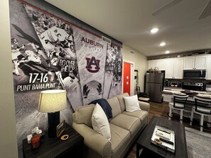 A living room with a couch, coffee table, lamp, and a wall decorated with Auburn football memorabilia. - Auburn U Campus condo! Near campus. Perfect for gamedays and family visits! (Auburn)