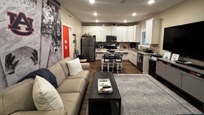 Una sala de estar con sofá, mesa de centro y televisor. Se observa una cocina con armarios blancos, refrigerador y microondas. Se ve un ventilador de techo.