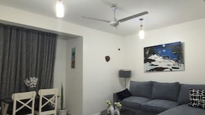 A living room with a grey sofa, a dining table with chairs, a ceiling fan, and a painting on the wall. - Greg black apartment - WITH AC/NETFLIX,HOTSHOWER (Mombasa)