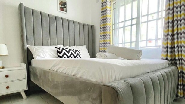 A bedroom with a grey upholstered bed, white bedding, a nightstand with a lamp, and a window with curtains. - Greg black apartment - WITH AC/NETFLIX,HOTSHOWER (Mombasa)