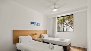 Una camera d'albergo con due letti, una ventola a soffitto, una finestra vista palme e un tavolino con una lampada.