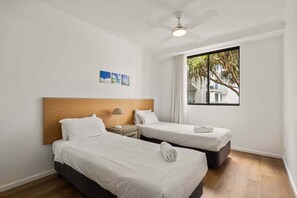 Ein Hotelzimmer mit zwei Betten, einem Deckenventilator, einem Fenster mit Blick auf Palmen und einem kleinen Tisch mit einer Lampe.