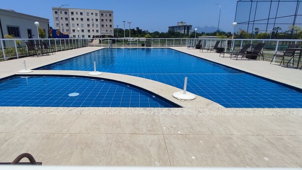Ein Schwimmbecken mit gefliestem Sonnendeck, Liegestühlen und Blick auf Gebäude im Hintergrund.