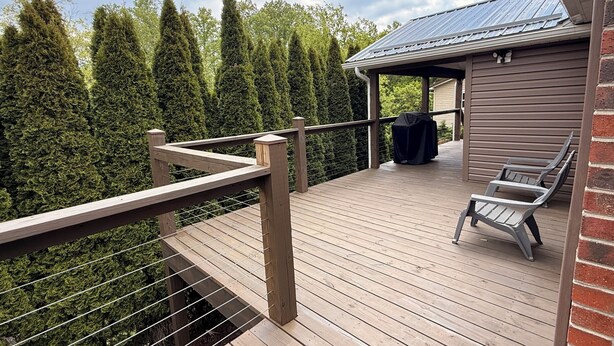 A wooden deck with a metal railing, a covered seating area with chairs, and a building with a metal roof. - Little Brick House - 10min from New River Gorge Bridge (Fayetteville)
