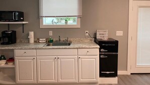 Una cocina con gabinetes blancos, microondas, cafetera, lavamanos y refrigerador.