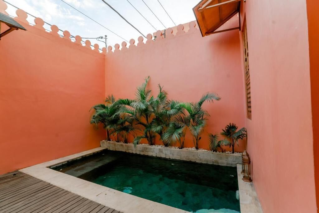 A small indoor pool with a wooden deck, surrounded by palm plants and a pink wall.