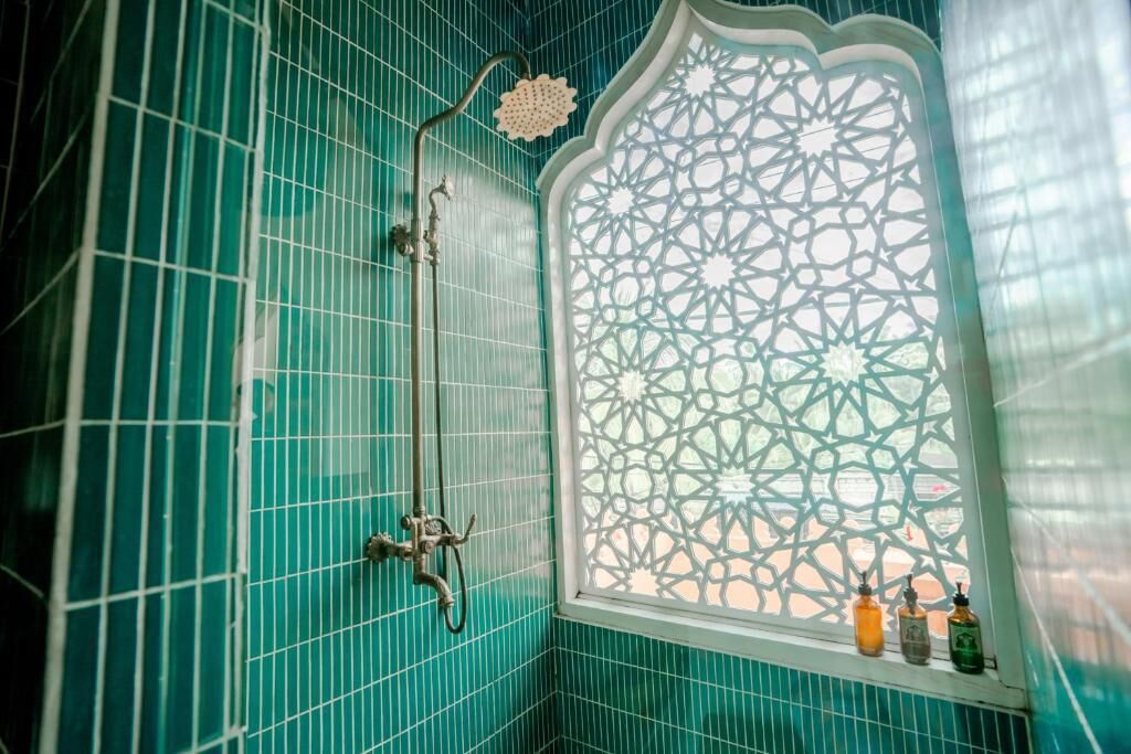 A bathroom with teal tiles, a shower with a curved design, and a window with intricate lattice work.
